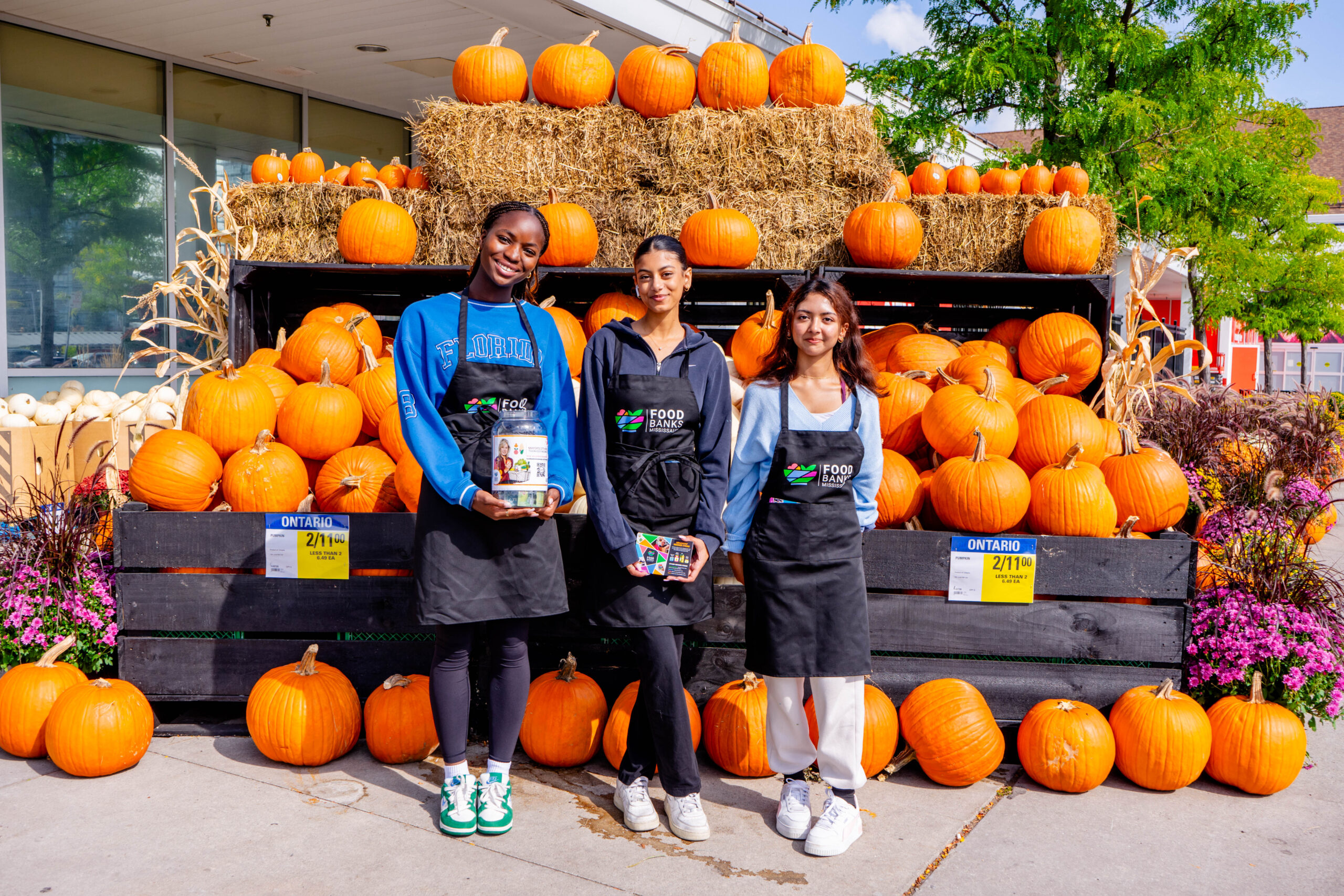 Three volunteers at a grocery store collecting donations for the Thanksgiving Drive standing in front of a display of pumpkins and hay.