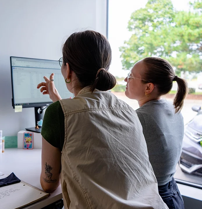 Two people at standing desk facing the computer, one person points at screen