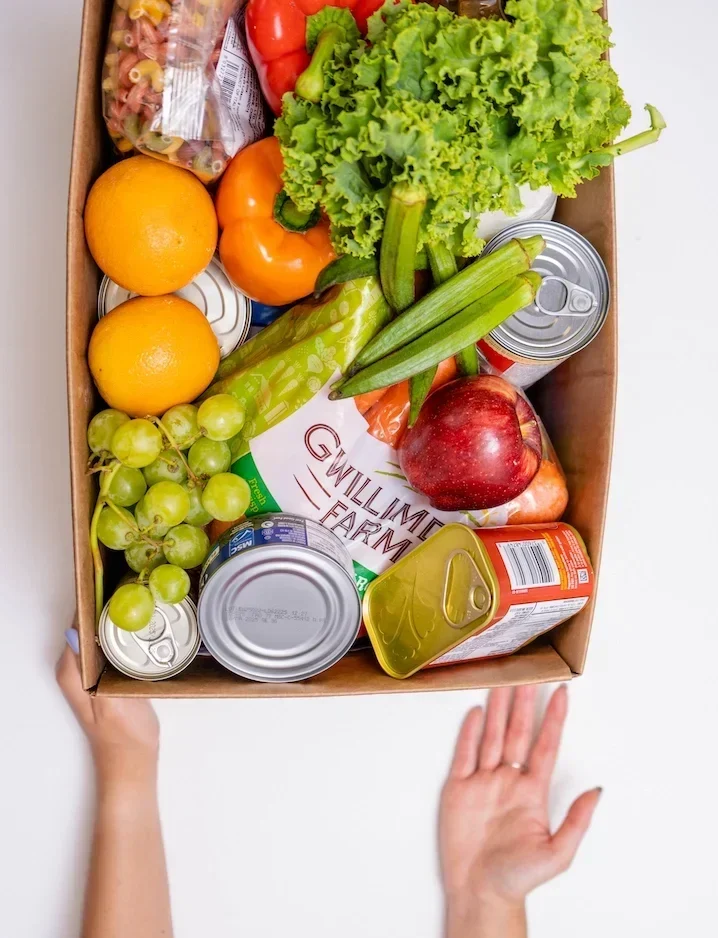 Hands presenting open box of food donations with fresh and shelf-stable foods