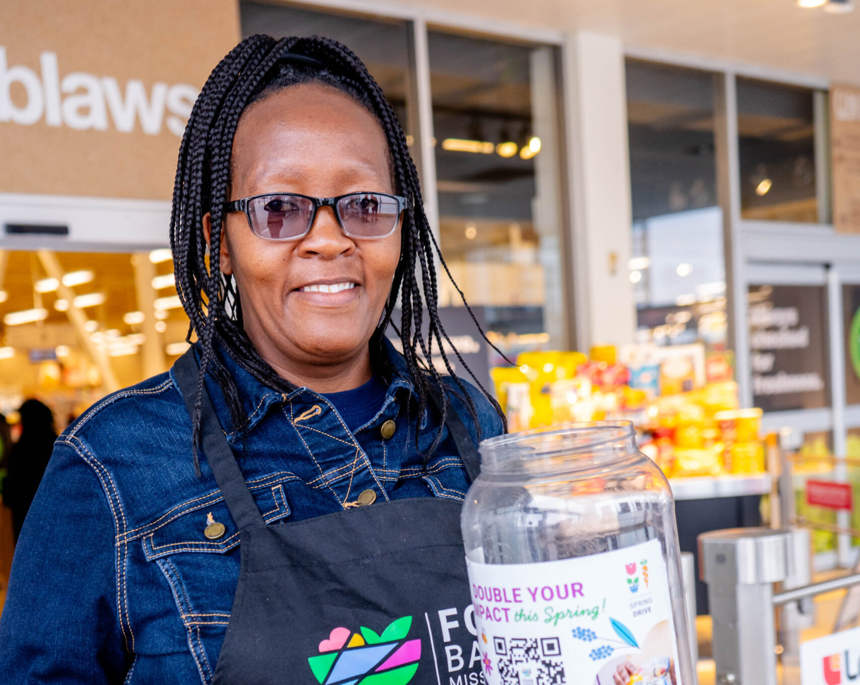 Volunteer in a jean jacket, wearing a black Food Banks Mississauga apron inside a grocery story holding a coin jar to collect donations.