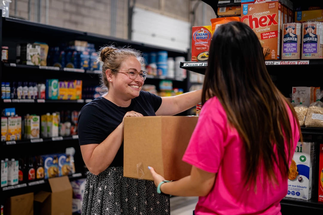 Food bank team member helping a volunteer pack food into a hamper in the Food Bank 2 Home Delivery Program area of the warehouse.