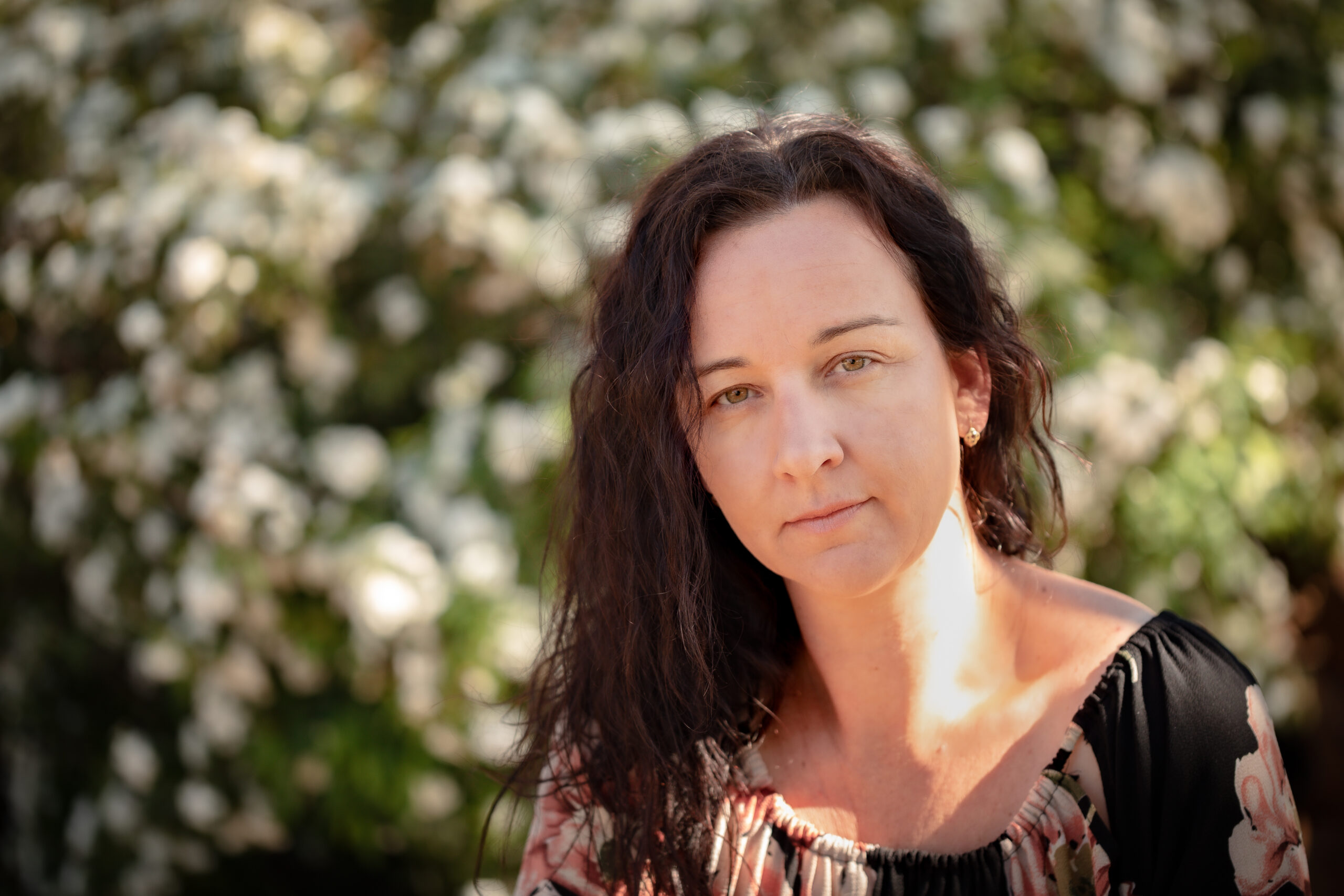 A caucasian female wearing a floral dress sitting in front of a wall of white flowers.