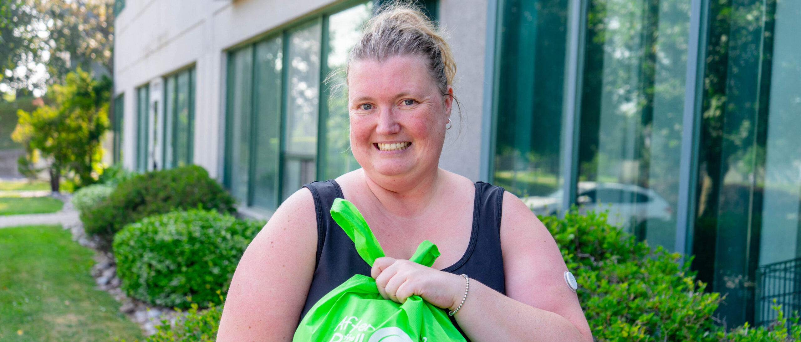 Food bank visitor with blonde hair in a ponytail, wearing a black tank top, holding a bright green bag with food, outside the building of a network food program.