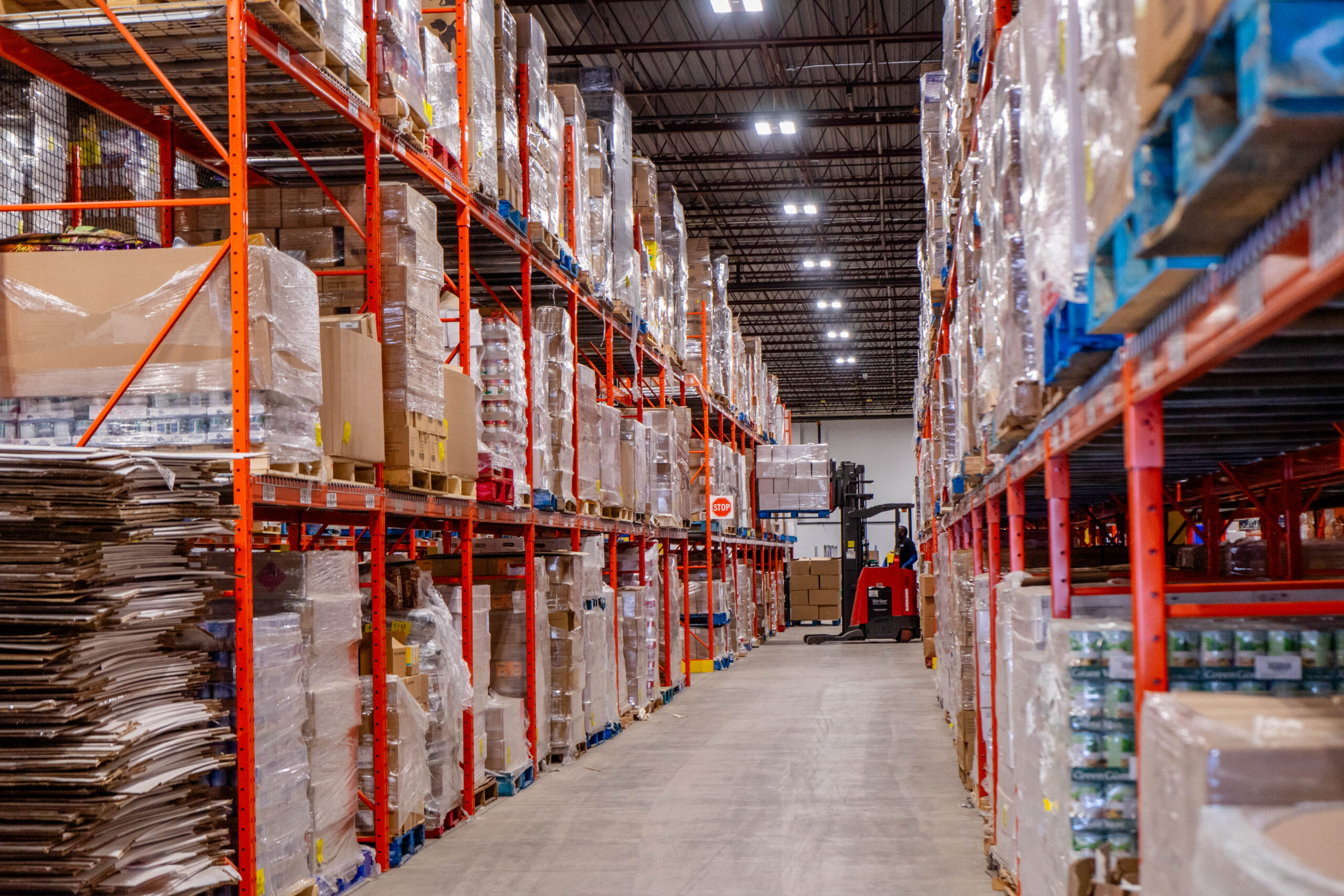 An aisle in the Food Banks Mississauga warehouse, showing racks stocked full of pallets of food with a warehouse operator using a forklit to lift a pallet onto the top rack.