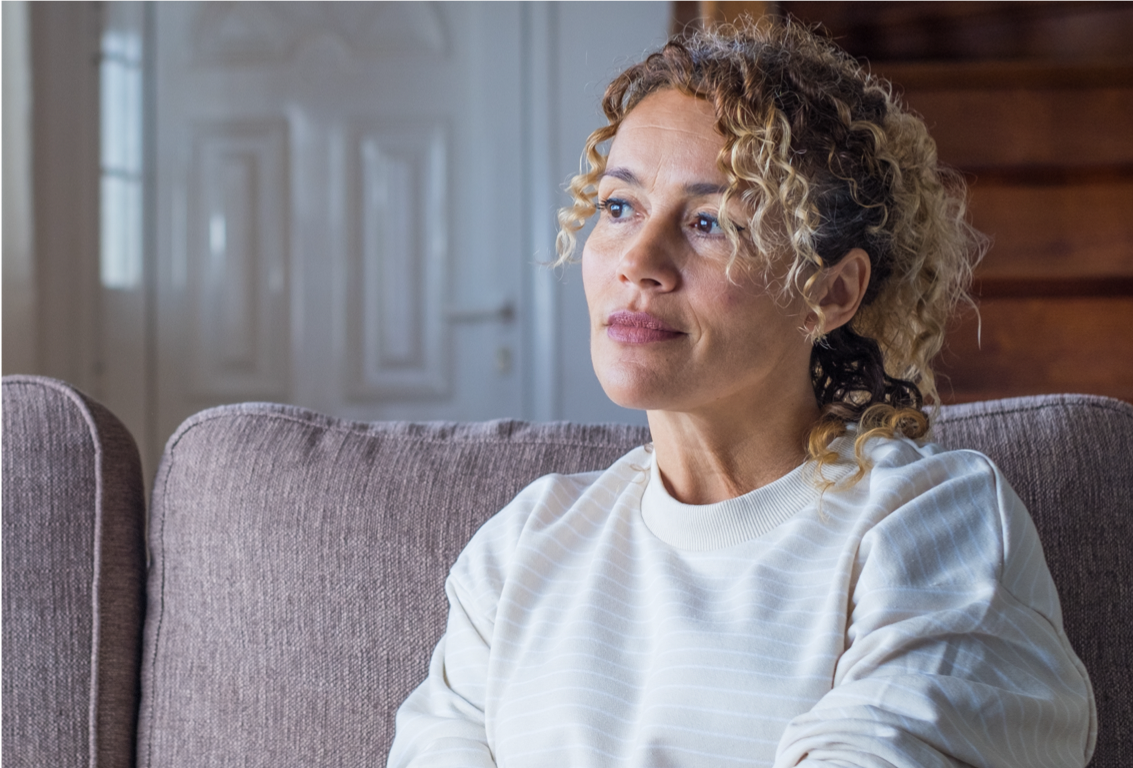 Woman with curly blonde hair in a white sweater, sitting on a couch in her home, looking off into the distance.
