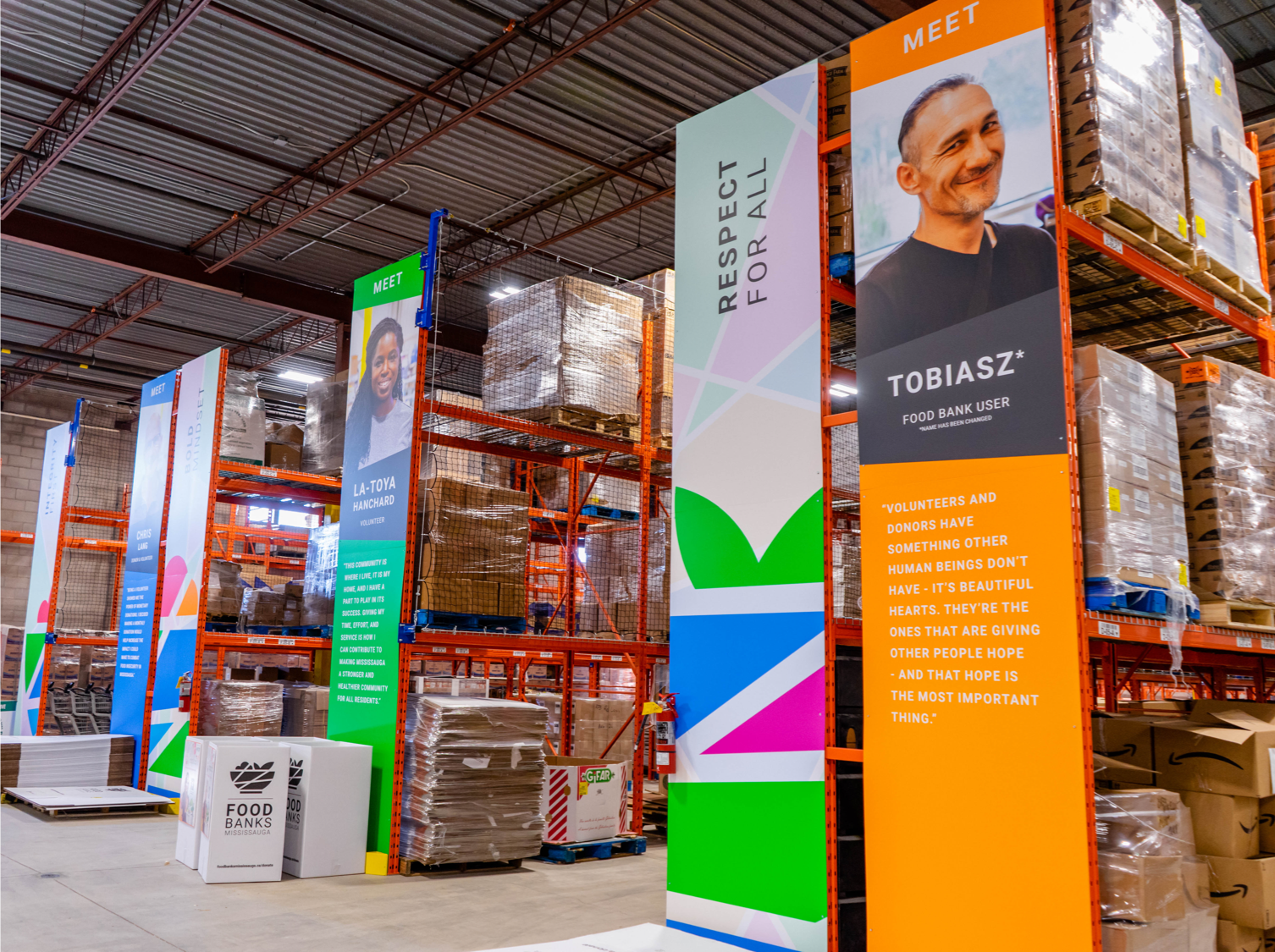 Interior of Food Banks Mississauga warehouse, showing racks full of pallets of food and colourful banners with a food bank visitor photo and quote on one banner.